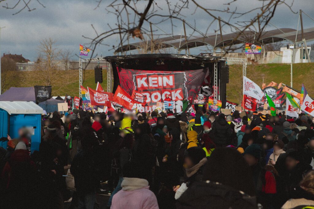 Große Menschenmenge mit Fahnen verschiedenster politischer Organisationen. Links ein Dixiklo, im Hintergrund prominent sichtbar eine Bühne mit einem großen Transparent mit der Aufschrift "Kein Bock auf Nazis". Dahinter sind Stadiontraversen auf einem Hügel zu sehen.