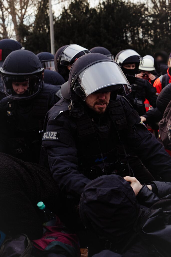 Zu sehen sind zahlreiche behelmte Polizist*innen. Im Hintergrund auch Rettungskräfte. In der Mitte des Bildes ist zu sehen wie ein Polizist gegen einen sitzenden Menschen welcher im Bild von Hinten zu sehen ist und eine schwarze Regenjacke trägt Gewalt anwendet.